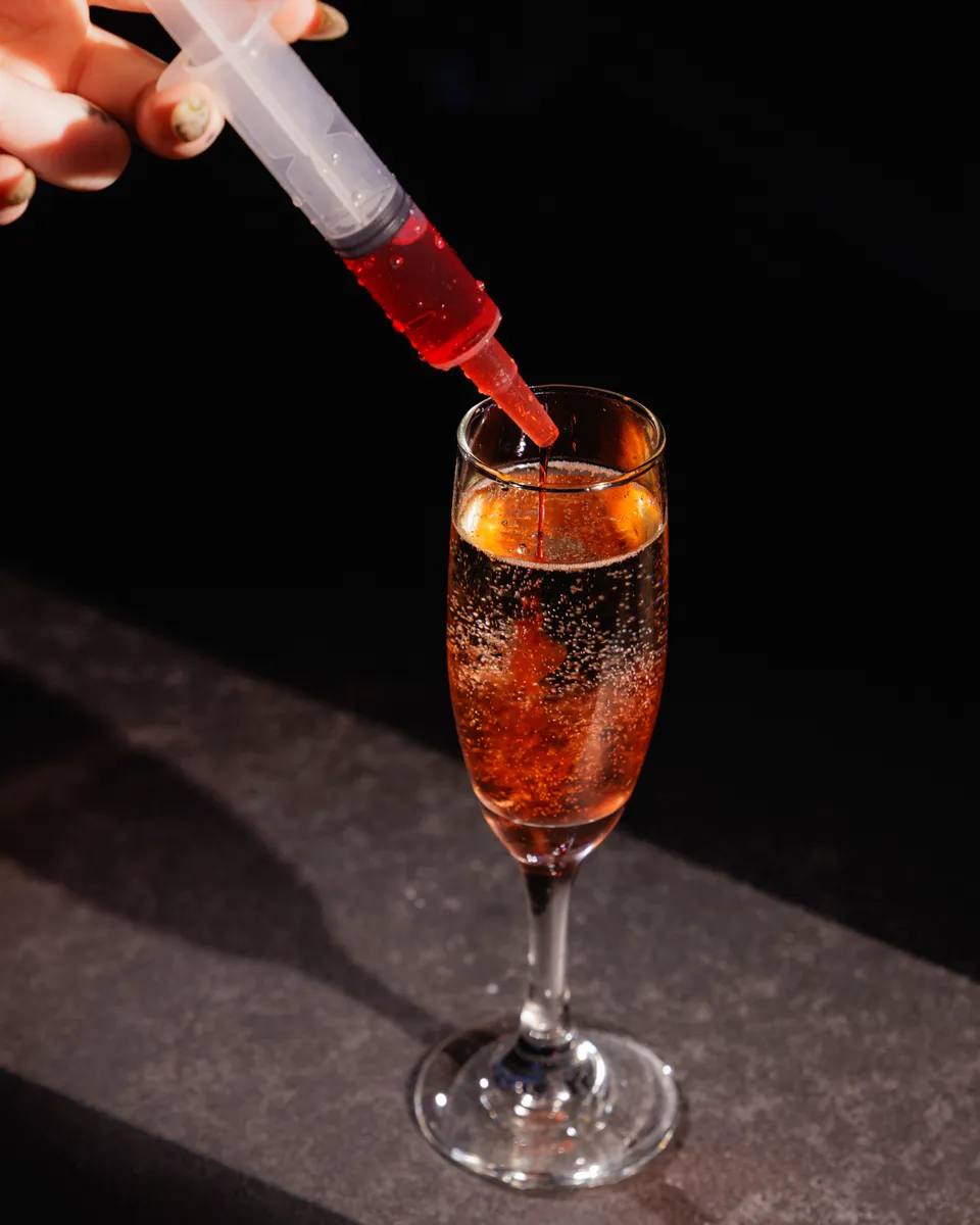 Red liqueur poured from a syringe into a sparkling champagne cocktail