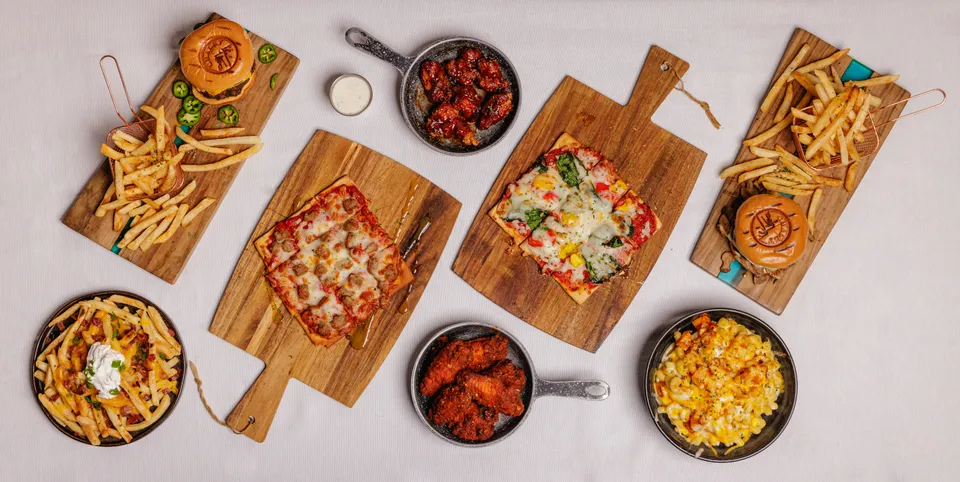 Overhead table spread of Detroit-style pizzas, wings, and sides on wooden boards