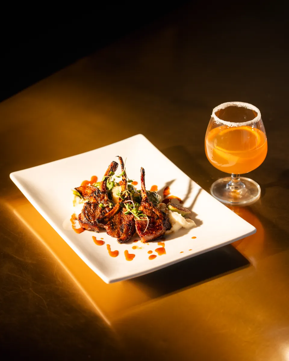 Plated lamb chops with a craft cocktail under warm spotlight